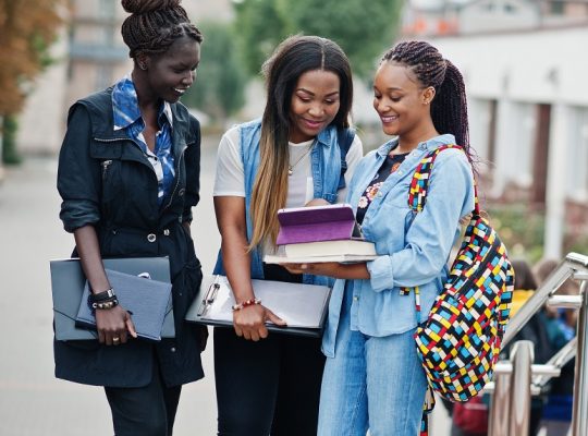 Three african students female posed with backpacks and school items on yard of university and look at tablet.